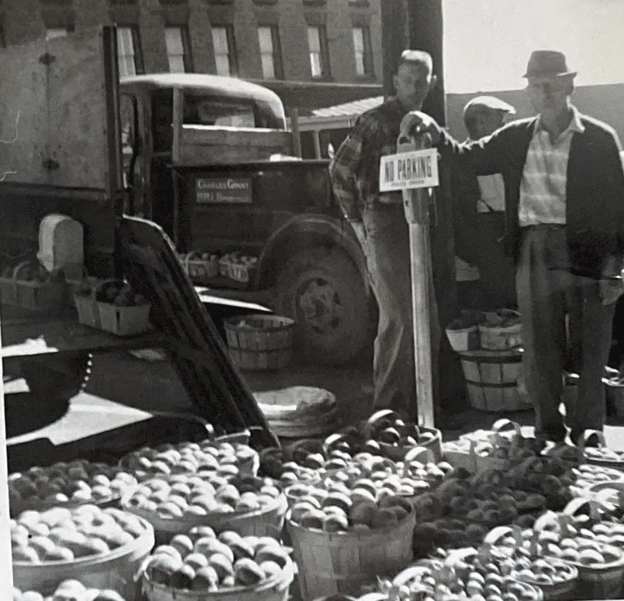 Men standing behind rows of apple baskets at a downtown market, with a truck parked behind them