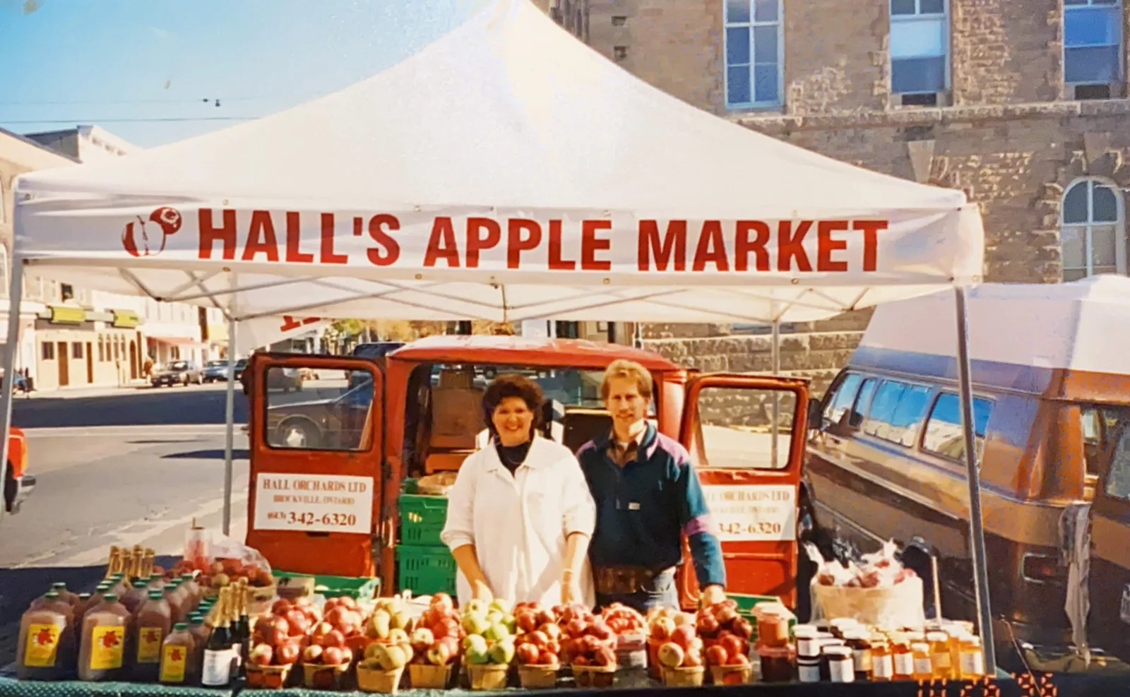 Kim and Chris Hall smiling behind a table of apple baskets, cider jugs and preserves under a white Hall’s Apple Market tent