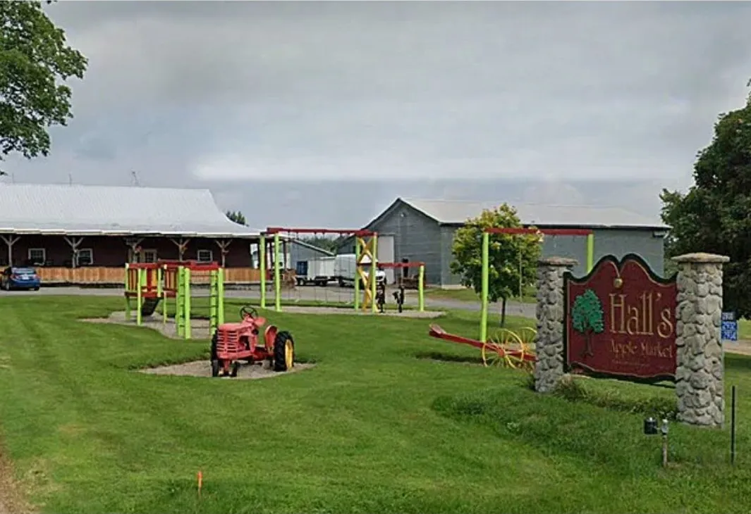 The Hall’s Apple Market sign by the road, with the market, play equipment and a vintage red tractor on the lawn behind it